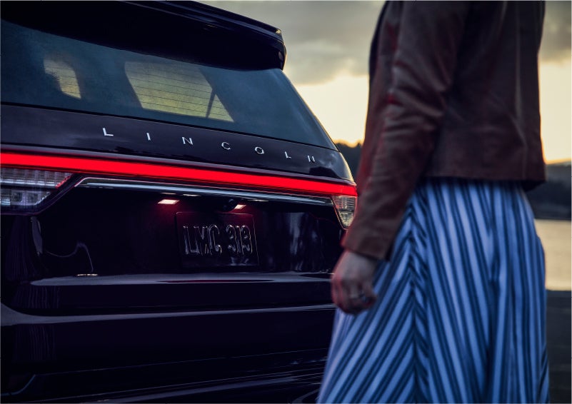 A person is shown near the rear of a 2023 Lincoln Aviator® SUV as the Lincoln Embrace illuminates the rear lights | Dorsch Lincoln in Green Bay WI