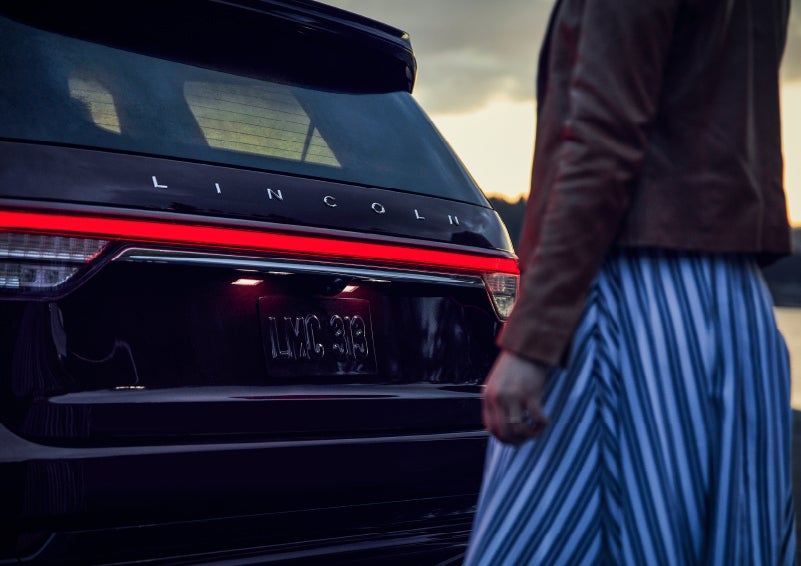 A person is shown near the rear of a 2024 Lincoln Aviator® SUV as the Lincoln Embrace illuminates the rear lights | Dorsch Lincoln in Green Bay WI