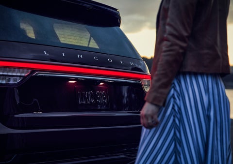 A person is shown near the rear of a 2024 Lincoln Aviator® SUV as the Lincoln Embrace illuminates the rear lights | Dorsch Lincoln in Green Bay WI